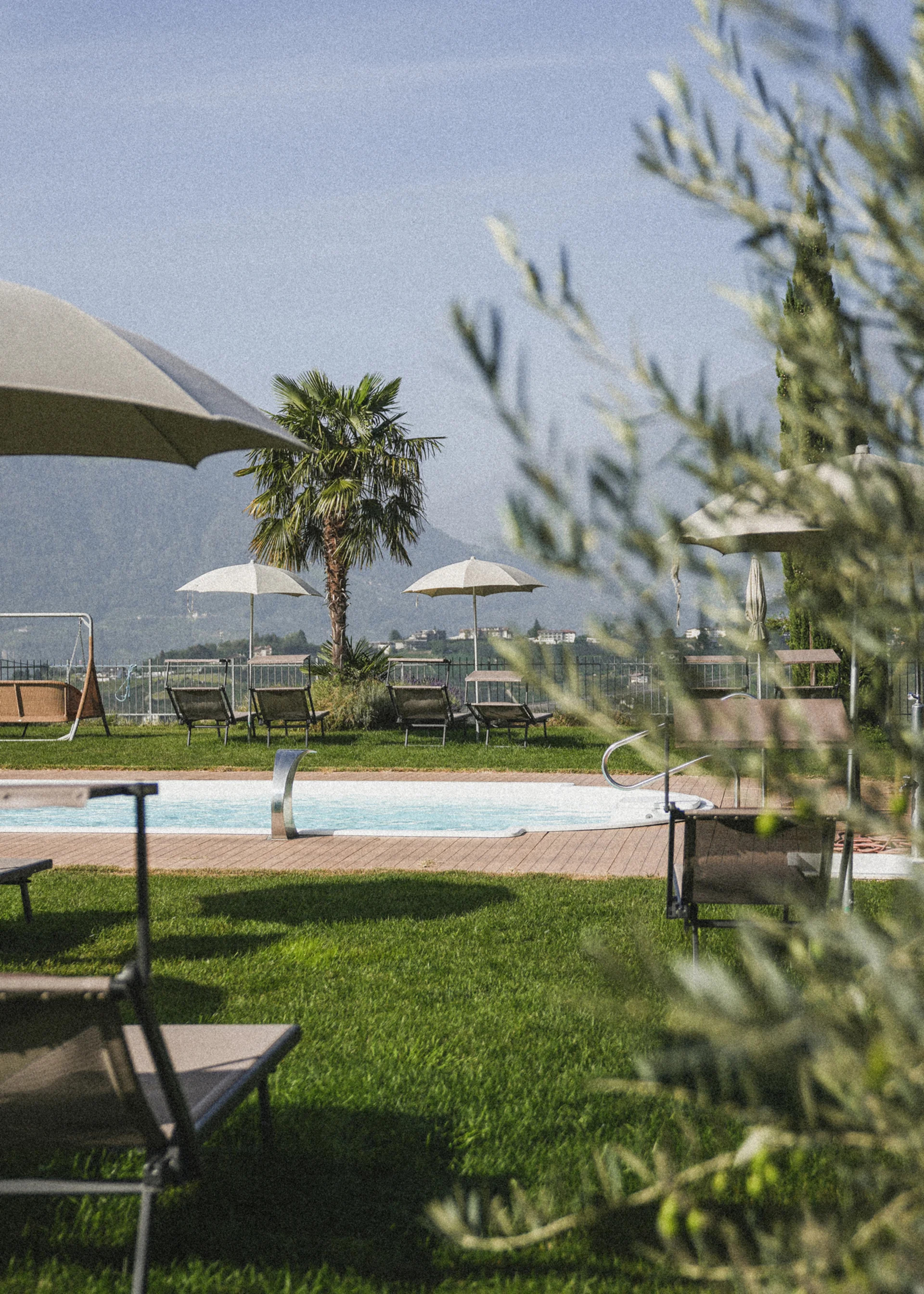 Euer Hotel in Schenna für Momente in #echtzeit Pool mit Liegestühlen und Sonnenschirmen in einer Gartenanlage mit Bergen im Hintergrund