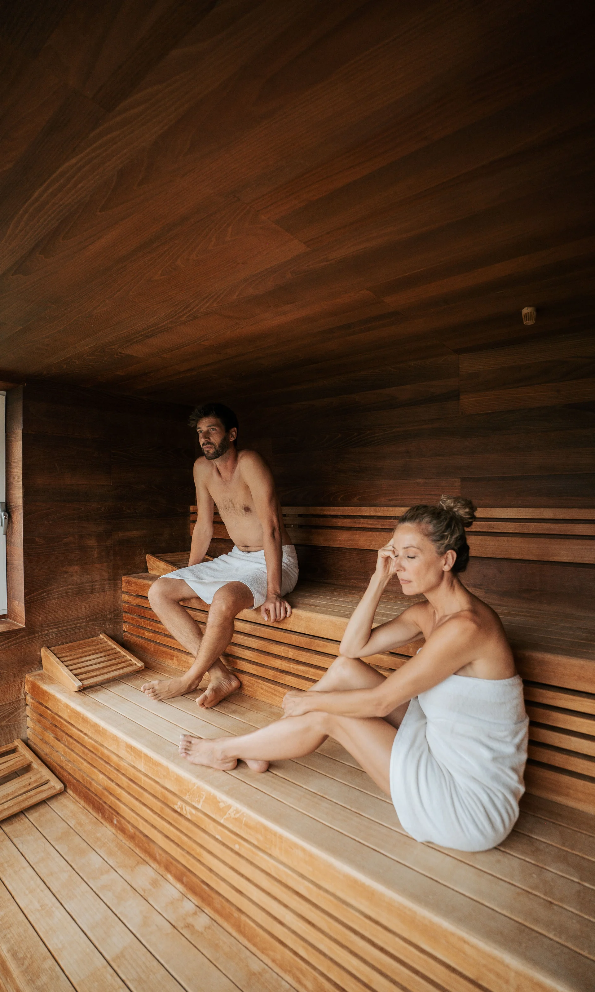 Euer Hotel in Schenna für Momente in #echtzeit Mann und Frau sitzen in einem Holzsauna-Raum mit weißen Handtüchern