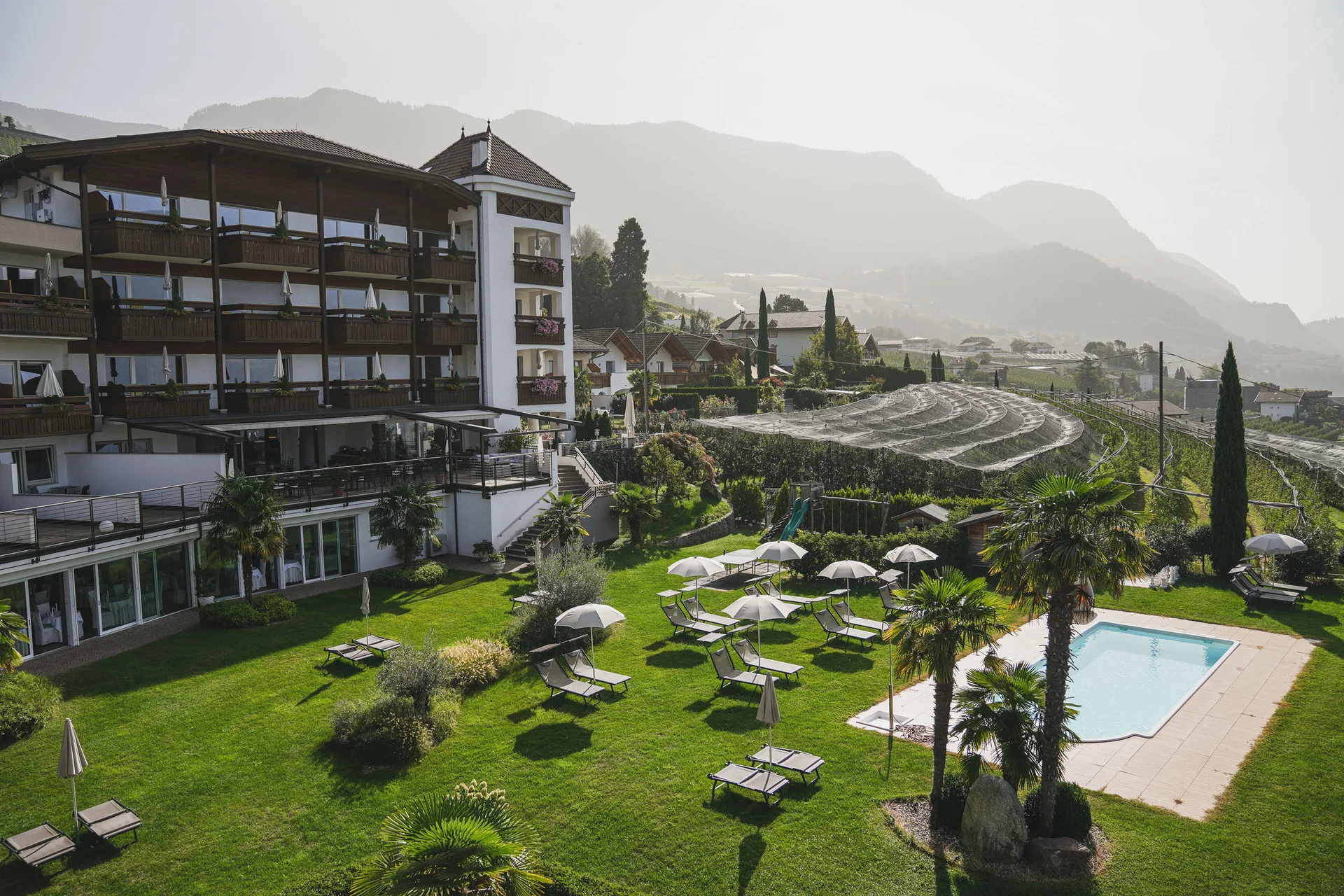 Euer Hotel in Schenna für Momente in #echtzeit Hotel mit Liegestühlen, Sonnenschirmen und Pool in einer grünen Berglandschaft